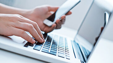 Bild einer Person, die mit der einen Hand auf einem Laptop tippt und in der anderen ein Smartphone hält