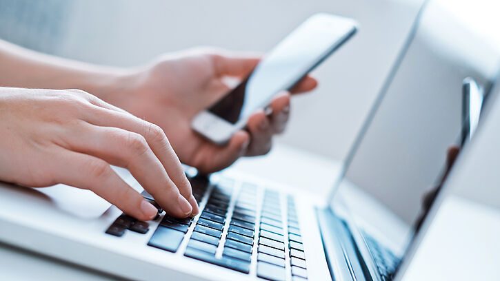 Foto zeigt, wie eine Person mit der einen Hand auf einer Laptop-Tastatur tippt und mit der anderen Hand ein Smartphone hält