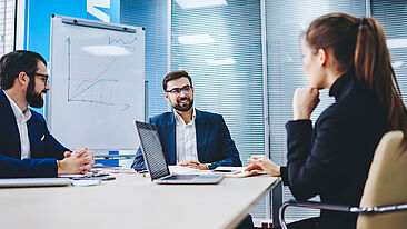 Drei Personen in Business-Kleidung sitzen um einen Tisch und unterhalten sich