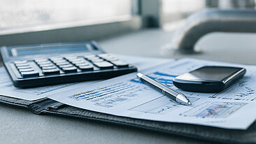 Bild zeigt Dokumente mit Auswertungen, einen Stift, ein Smartphone und einen Taschenrechner